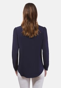 Navyblauwe blouse met lange mouwen, een soepele textuur, afgeronde zoom en geplooid schouderdetail. Gemaakt met witte jeans.