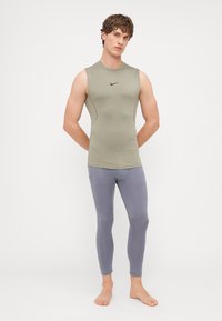 Oblikovaný bezrukávový olivovo zelený top s logom Nike, spárovaný s priliehavými šedými legínami s akcentmi a švami, model bosý.