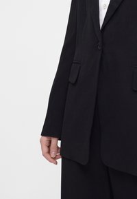Personne portant un blazer noir à un bouton avec poches à rabat sur une chemise blanche, le bras et la main détendus sur le côté.