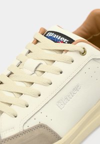 Baskets en cuir blanc avec des accents en daim beige, des lacets plats crème et une étiquette noire présentant le nom de la marque et un design de drapeau coloré.