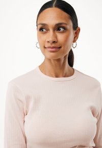 Frau mit zurückgebundenem dunklem Haar, die ein hellrosa geripptes Langarmoberteil und silberne Creolen trägt, leicht zur Seite blickend.