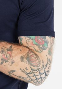 T-paita, tummansininen puuvillasta, lyhythihainen, esitetty käsivarren päällä, jossa on yksityiskohtaisia tatuointeja, kuten kukkia, sydämiä ja geometrisia kuvioita.