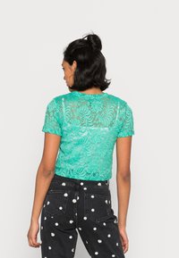 Top curto de renda turquesa com mangas curtas, apresentando padrões florais, textura semi-transparente, decote redondo e design cropped, combinado com jeans pretos com bolinhas.