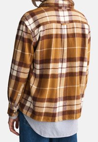 Camisa de franela a cuadros marrón y amarilla con cuello, puños con botones y tejido suave y texturizado. Lleva una camisa de rayas azules y blancas debajo.