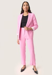Růžový přizpůsobený blazer a odpovídající kalhoty, s černým ribbovaným topem, s bílými pruhy na rukávech a černými špičatými balerínami.