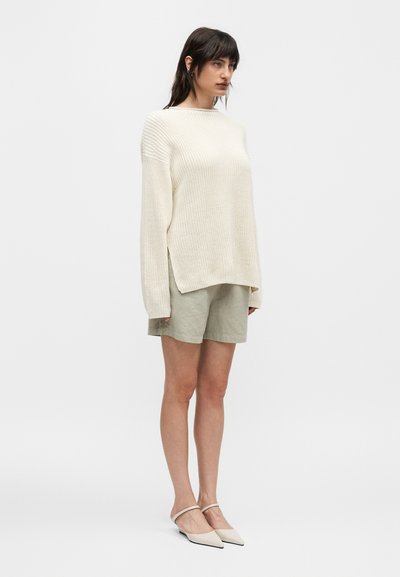 Γυναίκα που στέκεται στραμμένη ελαφρώς προς τα δεξιά, φοράει υπερμεγέθη πουλόβερ πλεκτό σε κρεμ χρώμα, ανοιχτό μπεζ σορτς και κρεμ μυτερές μπαλαρίνες με λουράκια στον αστράγαλο.