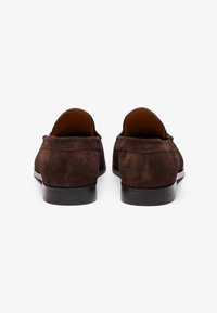 Braune Wildlederslipper mit matter Oberfläche, runder Zehenpartie und schwarzer Gummisohle. Sie zeichnen sich durch ein einfaches Design ohne sichtbare Beschläge oder Akzente aus.
