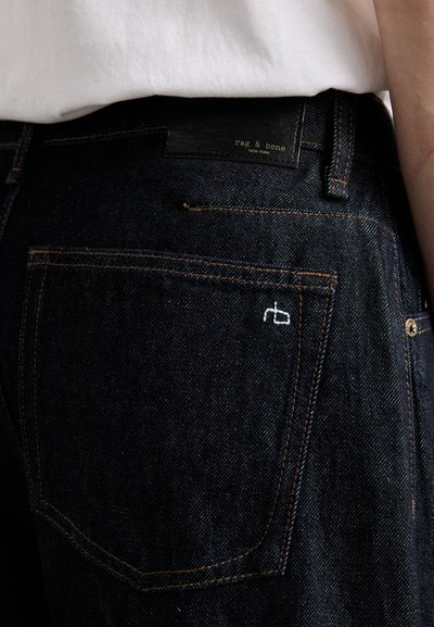 Dark denim jeans with orange stitching and a black leather waistband label. Features a white embroidered logo on the back pocket.