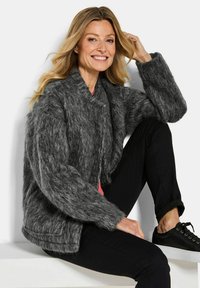 Grijze fluffy gebreide jas met een ronde kraag, sluiting aan de voorkant en brede mouwen, gecombineerd met zwarte broek en bijpassende sneakers.