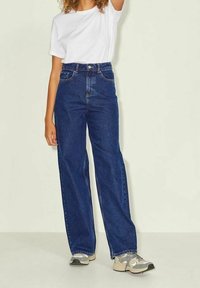 Höga, vida blå denimjeans med framfickor och kontrastsöm, i kombination med en vit t-shirt och grå sneakers.