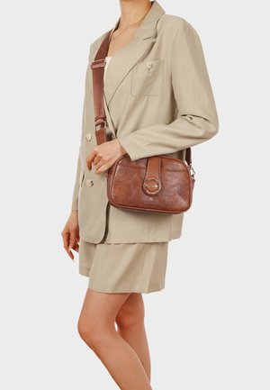 Donna che indossa un blazer beige e pantaloncini, con una borsa a tracolla in pelle marrone con chiusura circolare dorata.