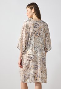 Kimono en dentelle avec motif paisley dans des tons beiges, dorés et gris. Coupe décontractée avec des manches larges, longueur mi-cuisse et texture légère.