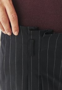Čierny látkový materiál s prúžkami a bielym švom, so vzorom vrecka. Ruka uchopila pás, čím ukazuje textúru látky.