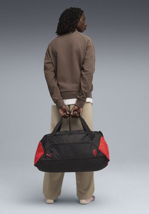 Sorte duffel bag med røde detaljer, udstyret med dobbelte håndtag og et logo. Fremstillet af slidstærkt stof med en glat tekstur og lynlås-lukninger.