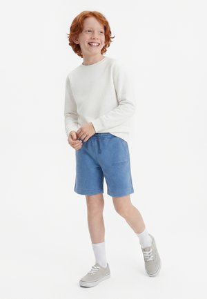 Rothaariger Junge in weißem Sweatshirt, blauer Shorts, weißen Socken und grauen Turnschuhen, lächelnd und vor einem einfarbigen Hintergrund stehend.