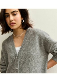 Cárdigan de lana gris con cuello en V, tejido texturizado y tres botones decorativos. Se lleva sobre una blusa blanca, con un ajuste suave y relajado.