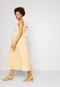 Vestido maxi em camadas de amarelo pálido com detalhe de laço no ombro. Combinado com sandálias de salto em malha bege. Textura suave, tecido leve.