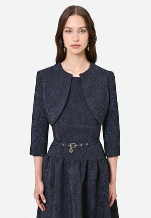Giovane donna con lunghi capelli castani indossa un abito blu scuro strutturato con una giacca corta abbinata e una sottile cintura con un piccolo charm.
