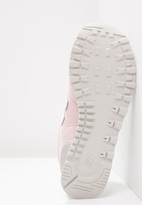 Semelle d'une basket blanche avec des crampons profonds et un logo, inclinée vers le haut contre une surface blanche en angle.