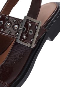 Sándalo de charol burdeos con un grueso tacón negro, adornado con tachuelas plateadas y una hebilla metálica rectangular en la correa.