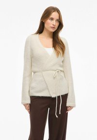 Krēmkrāsas pārklājuma kardigans no mīksta, pūkaina auduma. Tam ir gari piedurknes, jostas aizdare viduklī un ērtas, gaisīgas dizains.