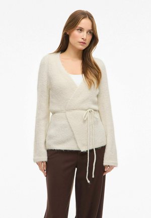Cardigan cache-cœur de couleur crème en tissu doux et moelleux. Il comporte des manches longues, une fermeture à nouer à la taille, et un design confortable et aéré.