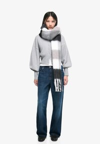 Scarf di lana grigio con strisce nere, bianche e grigie, indossato attorno al collo, abbinato a un cardigan in maglia grigia e a dei jeans in denim blu larghi.