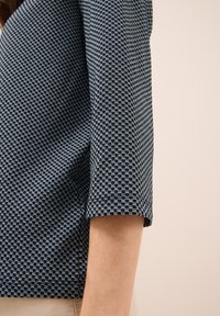 Mörkblå kortärmad topp med ett geometriskt mönster av sammanlänkade hexagoner i ljusare blått, med en strukturerad skärning och texturerad tyg.
