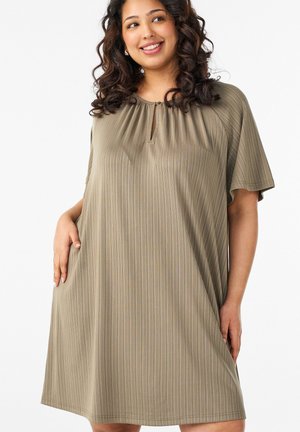 Glimlachende vrouw met krullend donker haar die een losse olijfgroene jurk met subtiele verticale strepen draagt, staand met één hand in de zak.