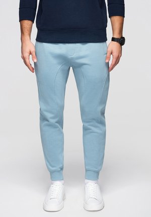 Mężczyzna ubrany w jasnoniebieskie spodnie dresowe, białe trampki, granatową bluzę i czarny zegarek na nadgarstku na jednolitym tle.