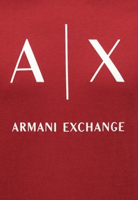 Κόκκινο ύφασμα με λευκό λογότυπο "A|X" πάνω από λευκό κείμενο "Armani Exchange".