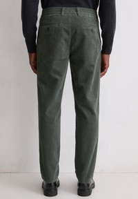 Groene corduroy broek met verticale strepen, met een aansluitende taille en twee achterzakken. Draagt met zwarte schoenen en een donkere trui.