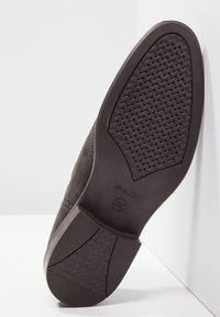 Semelle de chaussure en daim marron avec un dessous en caoutchouc texturé présentant un motif en chevrons et un logo embossé. Design plat, esthétique décontractée.