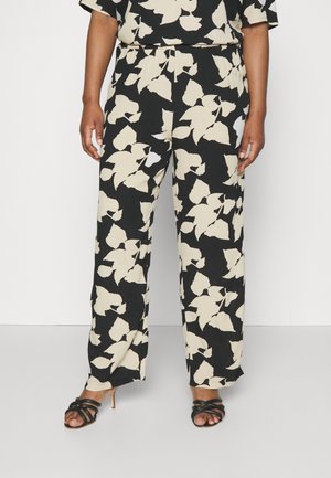 Persoon die losse zwarte broek draagt met grote beige bladerenprint en zwarte sandalen met hoge hak en bandjes, staand tegen een effen witte achtergrond.
