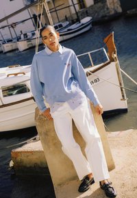 Ženska v svetlo modri majici s kapuco, belih hlačah in črnih čevljih se naslanja na pomol z ladjicami in vodo v ozadju.