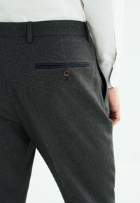 Pantalons grijs à carreaux avec une coupe ajustée, présentant une poche arrière avec un accent marine et un détail de bouton marron. Fabriqués en tissu texturé.