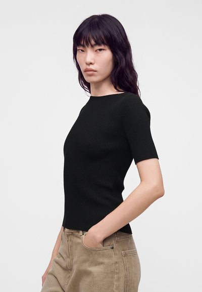 Femme aux longs cheveux foncés portant un haut noir côtelé à manches courtes et un pantalon beige, une main dans la poche, sur un fond clair uni.