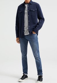 Granatowa koszula z guzikami noszona na podkoszulku w paski, zestawiona z niebieskimi dżinsami i czarnymi sneakersami. Posiada kieszenie na piersi i podwinięte mankiety.