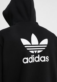 Person som bär en svart hoodie med en stor vit Adidas trefoil-logotyp och text på baksidan, vänd bort åt ett enkelt bakgrund.