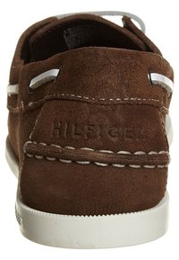 Los zapatos náuticos de ante marrón presentan cordones y costuras blancas, con el logo grabado en el talón. La suela es blanca, lo que contrasta con el material superior.