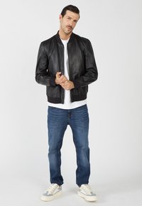 Zwarte leren bomberjack met ribbelmanchetten; gedragen over een wit T-shirt, gecombineerd met blauwe jeans en grijze sneakers. Gladde textuur.