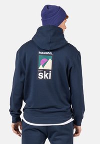 Felpa blu navy con finitura testurizzata, caratterizzata da un logo stampato sul retro che include forme geometriche e la parola "ski."