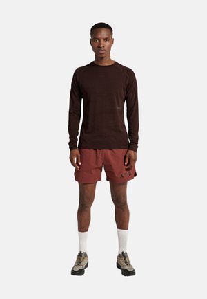 Man staat rechtop met zijn gezicht naar voren, gekleed in een donker langemouw shirt, bordeauxrode shorts met de tekst "ATW", witte sokken en beige wandelschoenen.