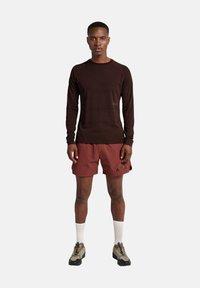 Mann steht mit dem Gesicht nach vorne, trägt ein dunkelgraues Langarmshirt, kastanienbraune Shorts mit "ATW"-Schriftzug, weiße Socken und beigetönige Wanderschuhe.