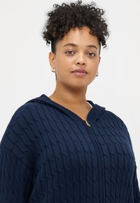 Ναυτικό μπλε φούτερ με φερμουάρ, φτιαγμένο από υφασμάτινη πλέξη υφής, με κουκούλα και χρυσό φερμουάρ, διαθέτοντας σχέδιο καλωδίου πάνω στο υλικό.