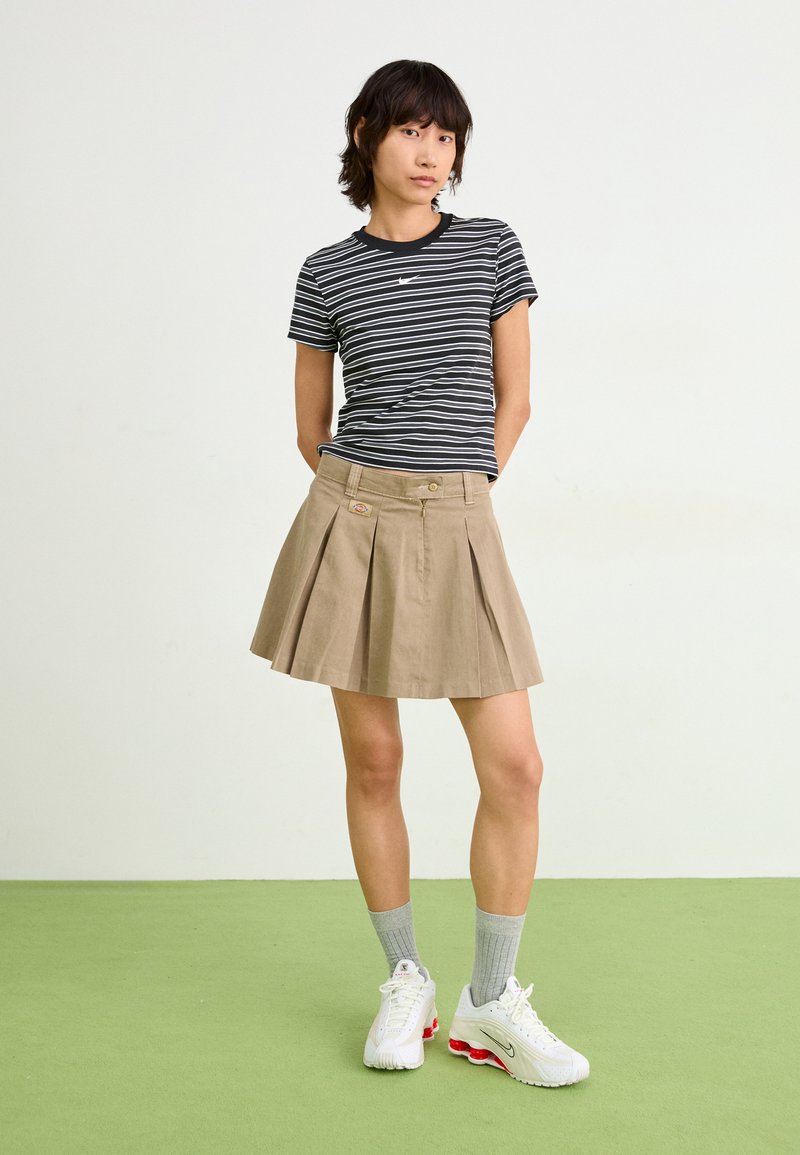 Czarny i biały w paski t-shirt w połączeniu z plisowaną beżową spódnicą. Modelka nosi szare skarpety i białe sneakersy z czerwonymi akcentami na zielonej podłodze.