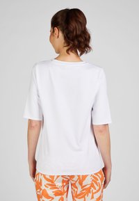 Weißes T-Shirt mit kurzen Ärmeln und rundem Halsauschnitt, glatte Textur und ohne sichtbare Muster. Von hinten vor einem neutralen Hintergrund gezeigt.