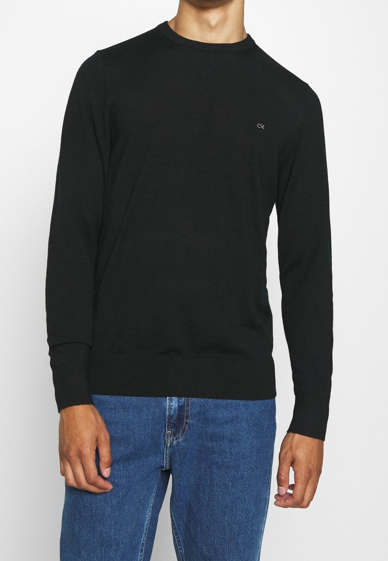 Homme portant un pull noir à manches longues avec un col rond et un petit logo "ck" sur la poitrine, ainsi qu'un jean bleu.