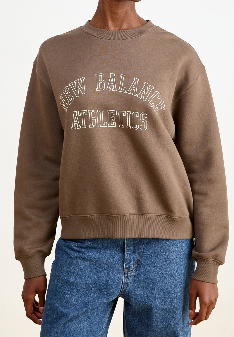 Brown sweatshirt with "NEW BALANCE ATHLETICS" text in cream. Features a round neckline and ribbed cuffs, paired with blue denim jeans.