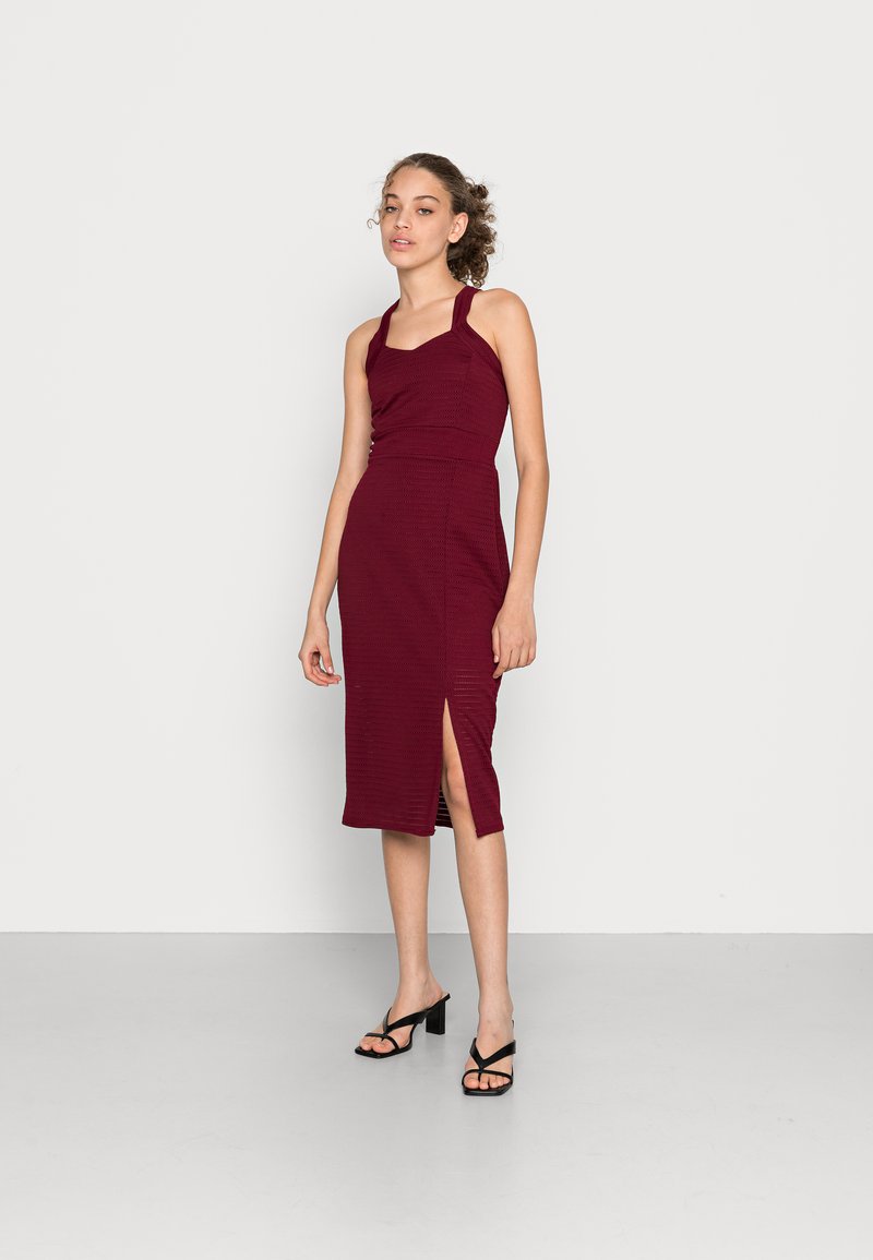 Vestido midi bordeaux com um design ajustado, textura canelada, decote halter e fenda lateral. Combinado com sandálias de salto altas pretas com tiras.